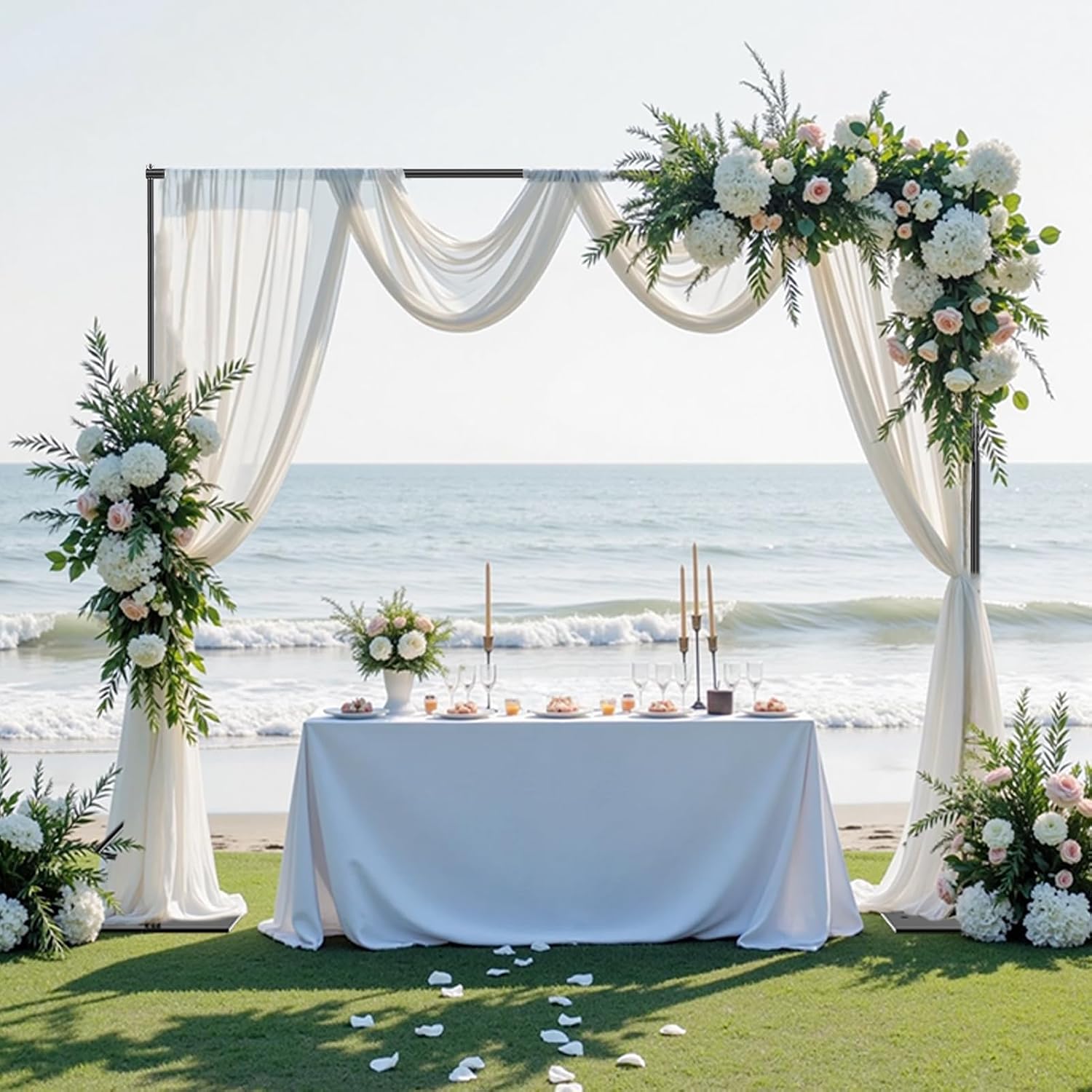 White drapes and floral arrangements on a flat-base backdrop stand behind a decorated table beside the sea.